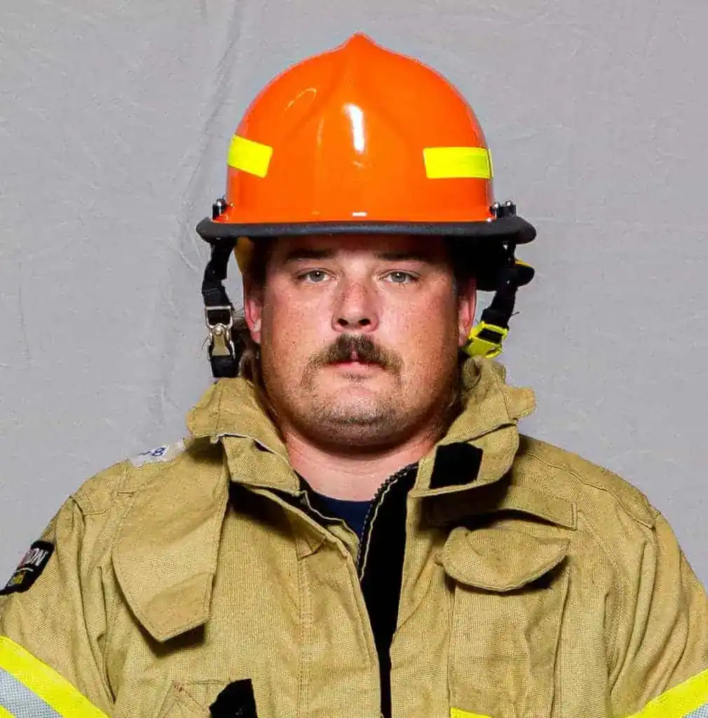 Justin Chambers, EMT with the Bahama Volunteer Fire Department, wearing full turnout gear and an orange helmet, facing forward in a formal portrait.