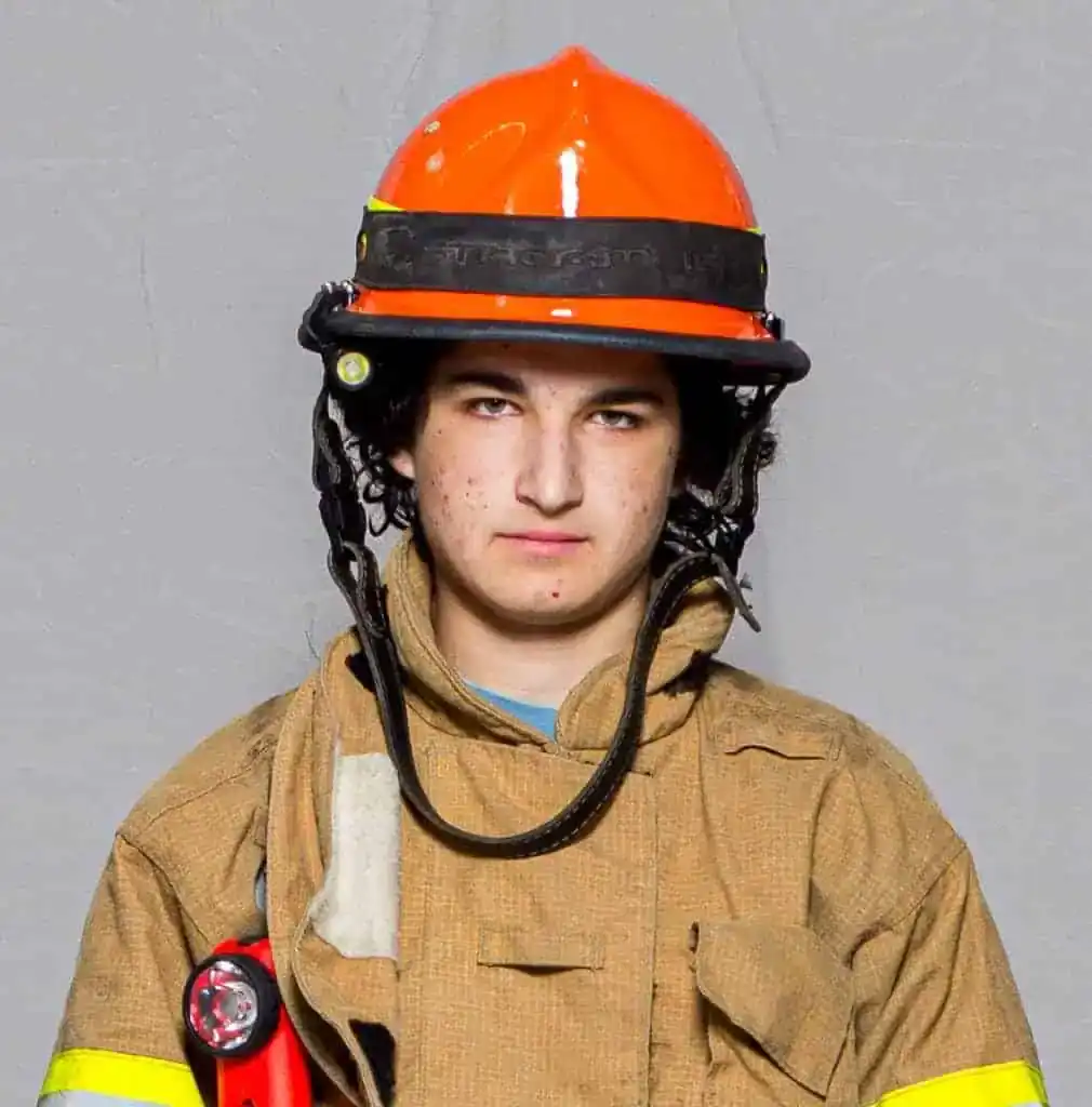 Colin Matthews, rookie with the Bahama Volunteer Fire Department, wearing full turnout gear and an orange helmet, facing forward in a formal portrait.