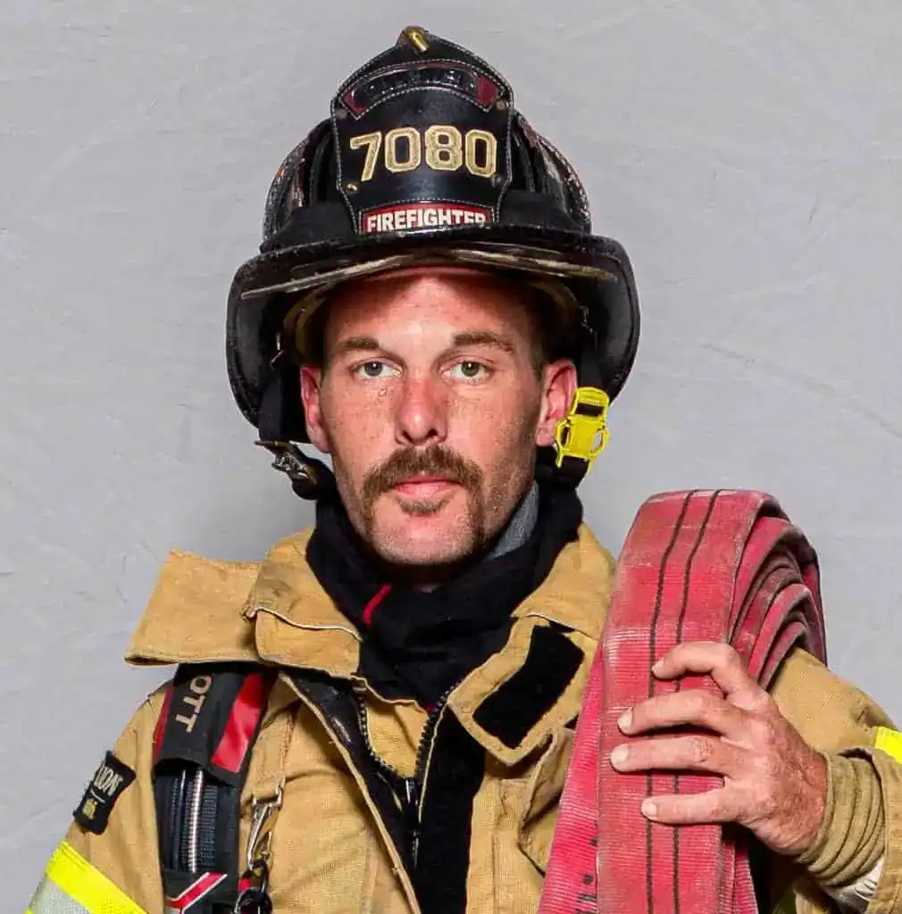 Ryan Baker, firefighter with the Bahama Volunteer Fire Department, in full turnout gear and a helmet labeled “7080 Firefighter,” holding a fire hose in a formal portrait.